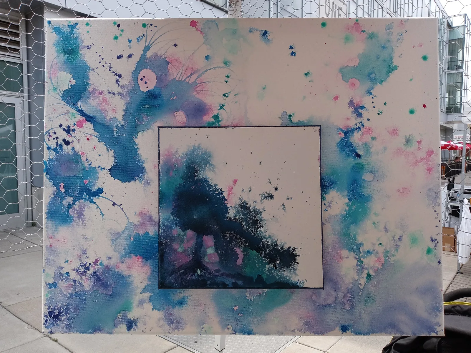 Large canvas held up outdoors: a dark inked tree on a small square panel set into a surrounding field of blue, pink and teal splatter.