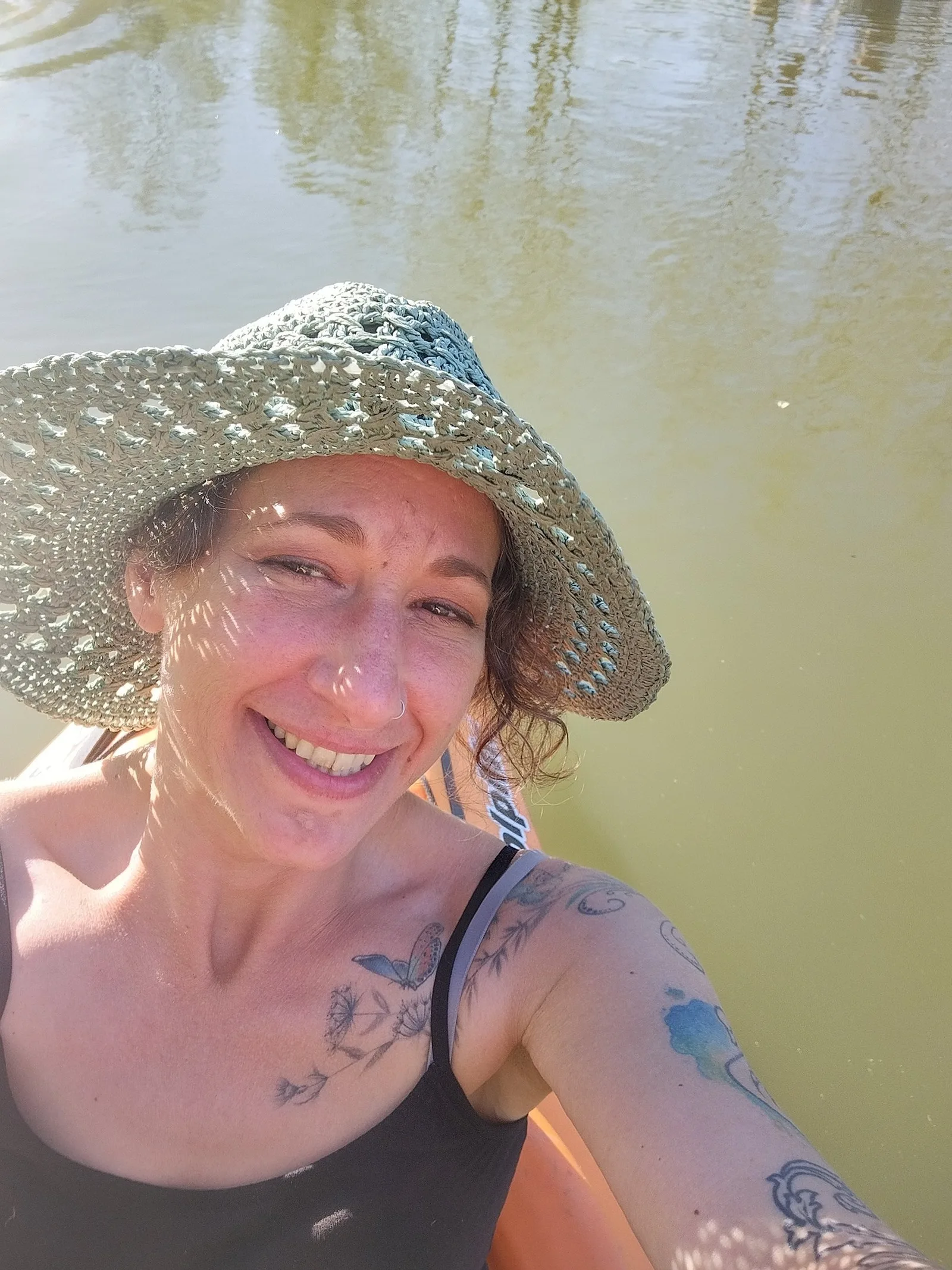 Angela outdoors on the water in a wide straw sun hat, smiling, dappled light across her face.