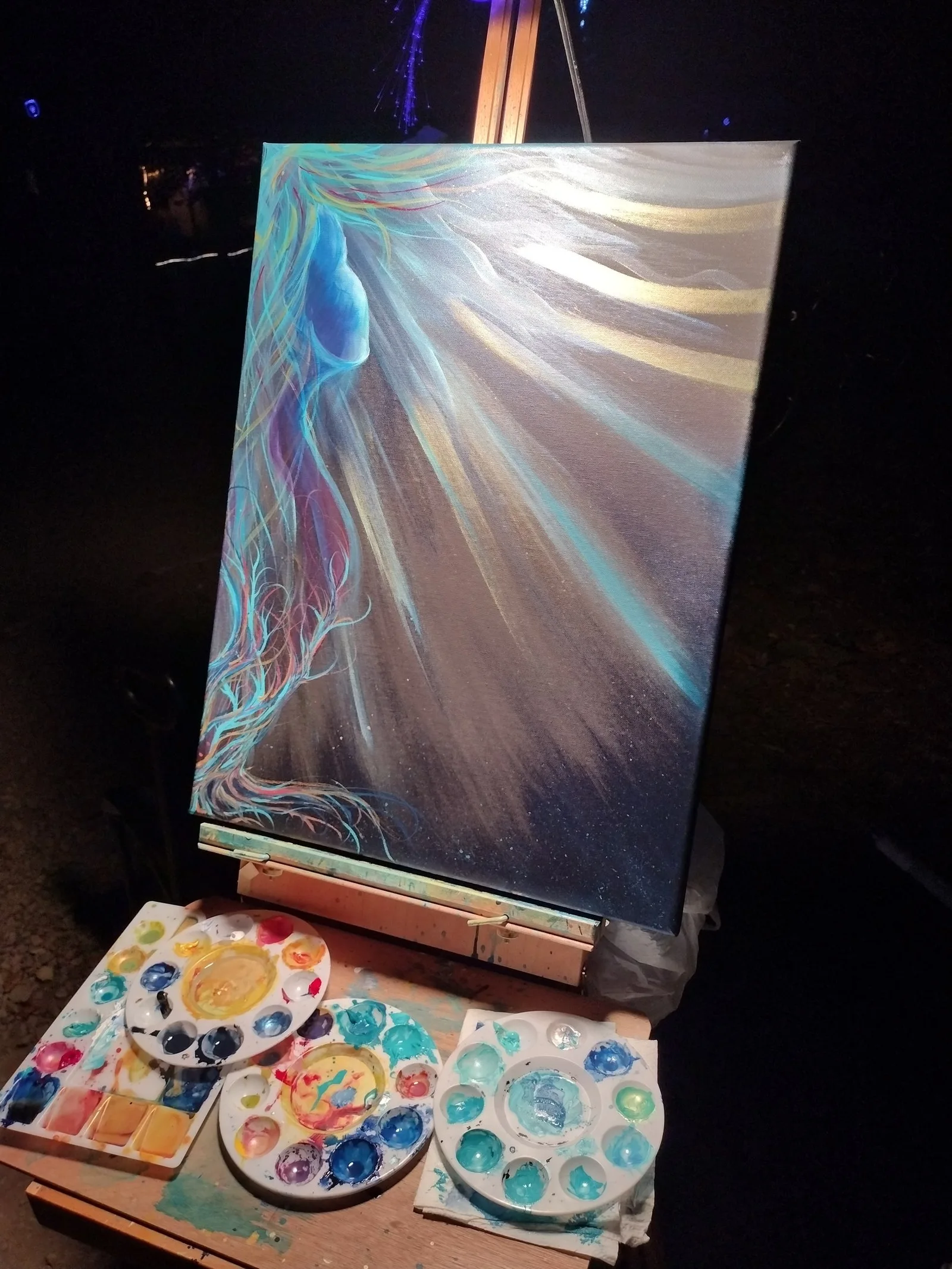 A canvas on an easel outdoors at night: a blue figure with long streaming hair beneath rays of gold and turquoise light, mixing palettes on the tray below.